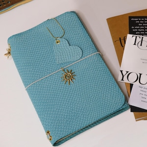 May include: A light blue journal with a textured, snake-skin pattern. The journal is secured with a white elastic band and adorned with a gold sun charm and a heart-shaped tag. Gold eyelets and a gold charm are visible.