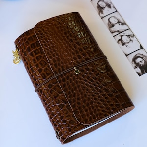 May include: A brown leather journal cover with a textured crocodile skin pattern. The cover is secured by a thin brown elastic band featuring a small green stone charm. A gold-toned metal charm hangs from the side, and a photo strip is nearby.
