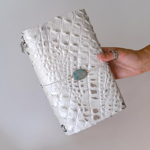 May include: A white journal with a textured crocodile skin pattern and a metallic silver finish. The cover is secured by a thin white elastic band featuring a central oval-shaped blue and gold stone accent. A small silver charm hangs from the side.