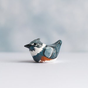 May include: Hand-painted ceramic bird figurine with blue, white, and orange details. The bird is perched on a white surface.