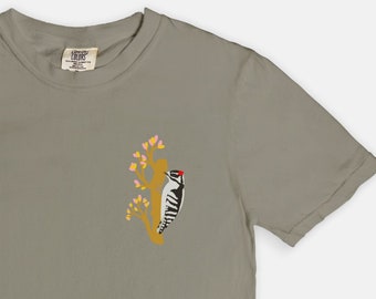 May include: A light gray t-shirt with a small embroidered image of a woodpecker on a branch with pink flowers.