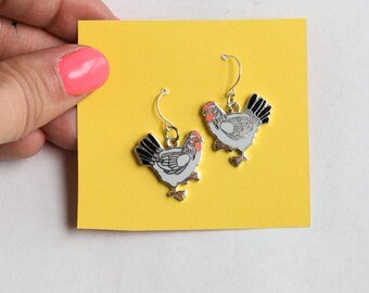 May include: A pair of silver chicken earrings on a yellow background. The chickens are white with black and orange details.
