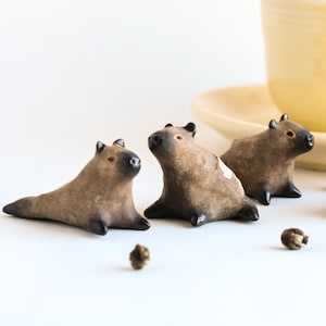 Puede incluir: Cuatro pequeñas figuritas de capibara de cerámica en marrón y negro, sentadas sobre una superficie blanca con hojas de té sueltas esparcidas a su alrededor. Una taza de té amarilla y un platillo están en el fondo.