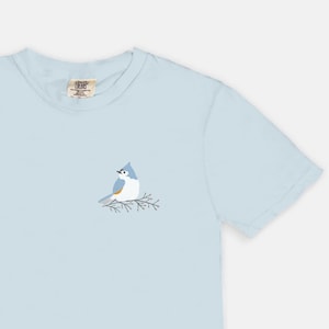 Könnte beinhalten: Ein hellblaues T-Shirt mit einem kleinen gestickten Bild eines blauen Vogels, der auf einem Ast sitzt.