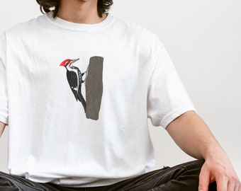 Puede incluir: Una camiseta blanca con una ilustración de un pájaro carpintero negro y blanco en la parte delantera. El pájaro carpintero está posado en un tronco de árbol.