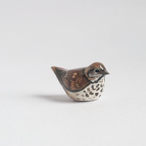 May include: A small, hand-painted ceramic bird figurine. The bird is brown and white with black markings and has a rounded body and a small beak.