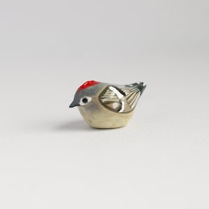Puede incluir: Pequeña figura de pájaro de cerámica. Tiene la cabeza roja, el cuerpo gris y marcas blancas y negras en las alas. El pájaro está sobre una superficie blanca. Mide aproximadamente 5 cm de largo.