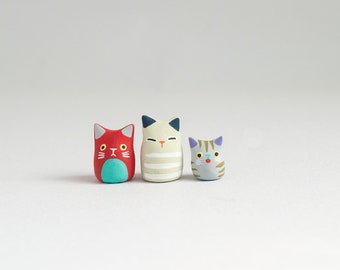 May include: Three small ceramic cat figurines. The cat on the left is red with a turquoise face. The cat in the middle is white with black ears and stripes. The cat on the right is gray with purple ears and a blue nose.