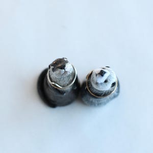 Pode incluir: Duas pequenas figuras de lontra cinzentas com anéis prateados ao redor do pescoço. As lontras têm detalhes em preto e branco nos rostos e estão posicionadas em uma superfície branca. As figuras são provavelmente feitas à mão.