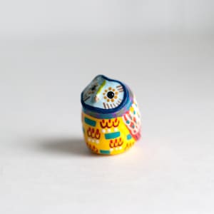 Könnte beinhalten: Eine kleine, handbemalte Keramik-Eulenfigur mit einem geometrischen Muster in Gelb, Blau, Rot und Grün. Die Eule hat einen blauen Kopf mit einem schwarzen Auge und einer weißen Pupille.