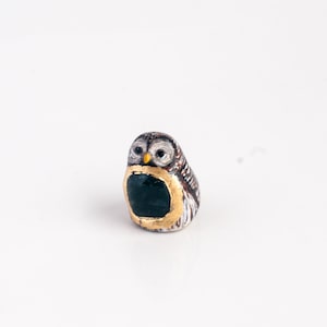 May include: A small ceramic owl figurine with brown, white, and black painted details. The owl has a gold ring around its body and a green painted chest.