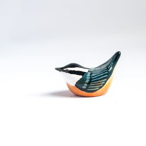 Op de afbeelding: Een klein keramiek vogeltje geverfd in tinten oranje, zwart, wit en blauw. Het vogeltje zit op een wit oppervlak.