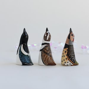 May include: Three ceramic figurines of birds, each painted with realistic details. The birds are a magpie, a quail, and a yellow-bellied sapsucker. The figurines are all facing upwards, as if they are singing.