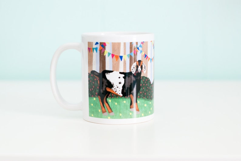 May include: White ceramic mug with a colorful illustration of a spotted goat standing in a field with trees and bunting in the background.