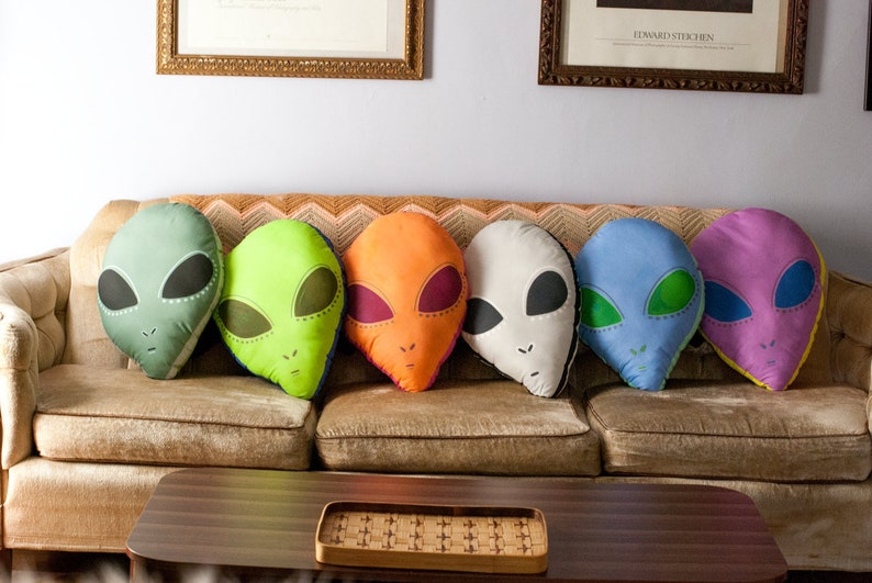 May include: Six colorful alien-shaped throw pillows on a brown couch. The pillows are green, orange, white, blue, and purple.