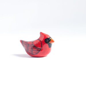 Peut inclure: Petite figurine de cardinal rouge avec des d&eacute;tails noirs et un bec orange. L'oiseau pr&eacute;sente un motif d'ailes d&eacute;taill&eacute; et une cr&ecirc;te. La figurine est pos&eacute;e sur un fond blanc, mettant en valeur les couleurs vives et le savoir-faire.