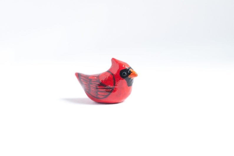 Peut inclure: Petite figurine de cardinal rouge avec des d&eacute;tails noirs et un bec orange. L'oiseau pr&eacute;sente un motif d'ailes d&eacute;taill&eacute; et une cr&ecirc;te. La figurine est pos&eacute;e sur un fond blanc, mettant en valeur les couleurs vives et le savoir-faire.