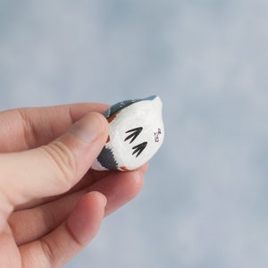 May include: A small, hand-painted ceramic figurine of a bird with blue, white, and brown markings. The bird is facing away from the camera and has a small, black inscription on its back.