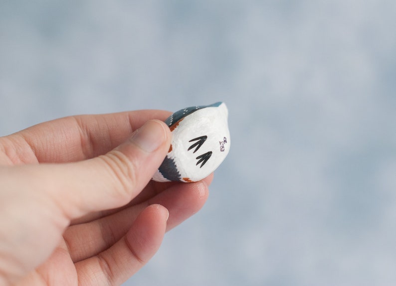 May include: A small, hand-painted ceramic figurine of a bird with blue, white, and brown markings. The bird is facing away from the camera and has a small, black inscription on its back.