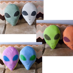May include: Six alien-shaped pillows in various colors, including green, white, orange, blue, and purple. The pillows are all on a brown and white patterned couch.