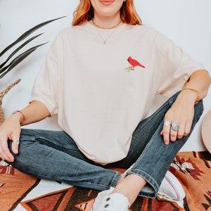 May include: A woman wearing a light beige t-shirt with a red cardinal embroidered on the front. She is sitting on a patterned rug with her legs crossed and wearing blue jeans and white trainers.