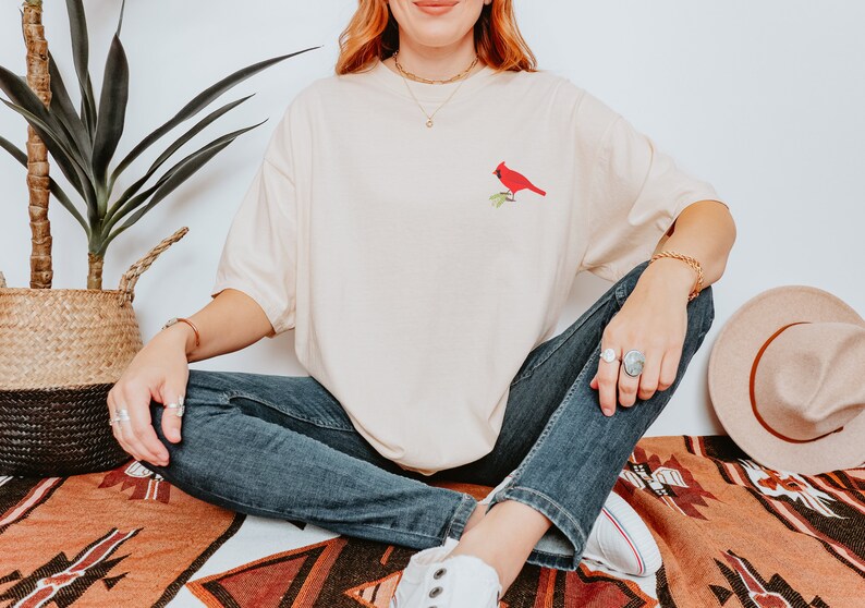 May include: A woman wearing a light beige t-shirt with a red cardinal embroidered on the front. She is sitting on a patterned rug with her legs crossed and wearing blue jeans and white trainers.