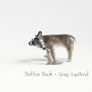 May include: A small, grey, ceramic squirrel figurine is being nuzzled by a larger, grey and brown, ceramic deer figurine. The deer has a black nose and black antlers. The figurines are on a white background. The text "Button Buck + Gray Squirrel" is below the figurines.