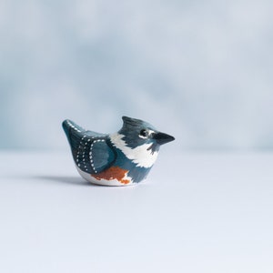 May include: Hand-painted ceramic bird figurine with blue, white, and orange details. The bird is perched on a white surface.