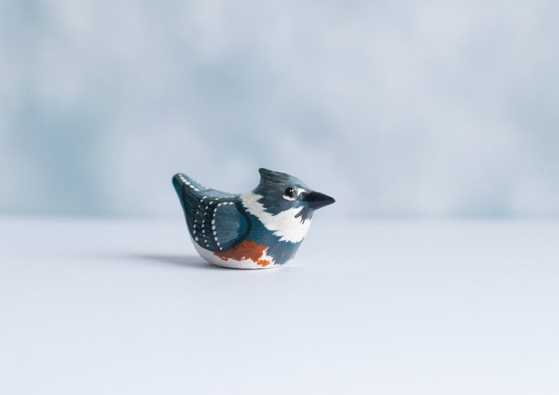 May include: Hand-painted ceramic bird figurine with blue, white, and orange details. The bird is perched on a white surface.