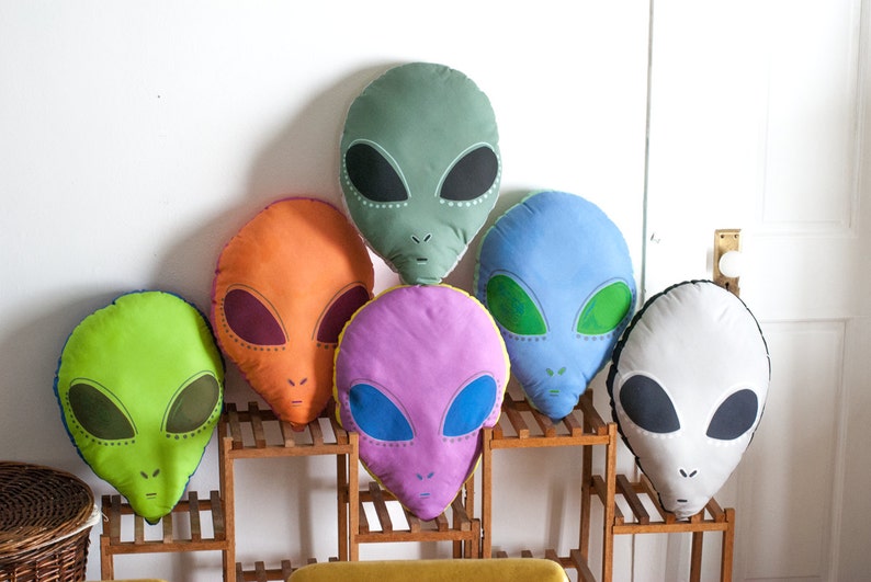 May include: Six colorful alien-shaped pillows with embroidered eyes and mouths. The pillows are arranged in a row on a wooden shelf.