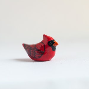 Peut inclure: Petite figurine de cardinal en c&eacute;ramique. L'oiseau est principalement rouge avec des d&eacute;tails noirs sur les ailes et le visage. Il a un petit bec orange, sur fond blanc.