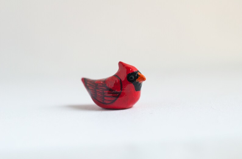 Peut inclure: Petite figurine de cardinal en c&eacute;ramique. L'oiseau est principalement rouge avec des d&eacute;tails noirs sur les ailes et le visage. Il a un petit bec orange, sur fond blanc.