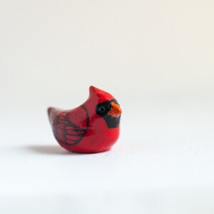 Peut inclure: Petite figurine de cardinal rouge faite &agrave; la main, avec des accents noirs et orange. L'oiseau a une cr&ecirc;te pro&eacute;minente et des marques d'ailes d&eacute;taill&eacute;es. La figurine est pos&eacute;e sur un fond blanc uni, mettant en valeur ses couleurs vives et son design.