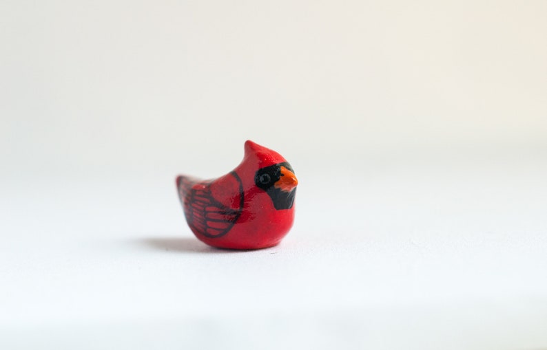 Peut inclure: Petite figurine de cardinal rouge faite &agrave; la main, avec des accents noirs et orange. L'oiseau a une cr&ecirc;te pro&eacute;minente et des marques d'ailes d&eacute;taill&eacute;es. La figurine est pos&eacute;e sur un fond blanc uni, mettant en valeur ses couleurs vives et son design.