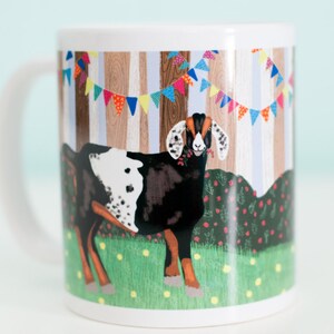 May include: White ceramic mug with a colorful illustration of a black and white goat standing in a field with a wooden fence and colorful bunting behind it.