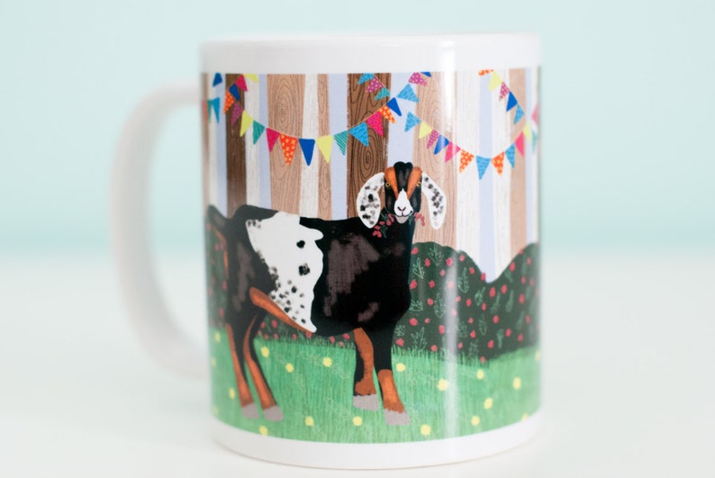 May include: White ceramic mug with a colorful illustration of a black and white goat standing in a field with a wooden fence and colorful bunting behind it.