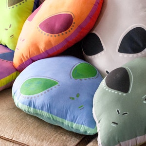 May include: Five colorful alien-themed throw pillows with different colored eyes and mouths. The pillows are made of soft fabric and have a variety of colors, including green, blue, pink, orange, and gray.