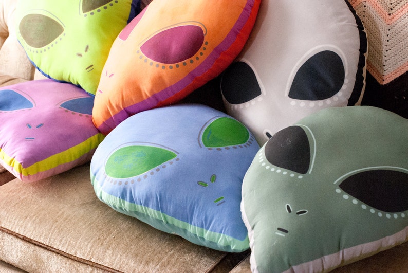 May include: Five colorful alien-themed throw pillows with different colored eyes and mouths. The pillows are made of soft fabric and have a variety of colors, including green, blue, pink, orange, and gray.