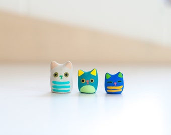 May include: Three small ceramic cat figurines, one white with blue stripes, one green with blue, and one blue with yellow stripes.