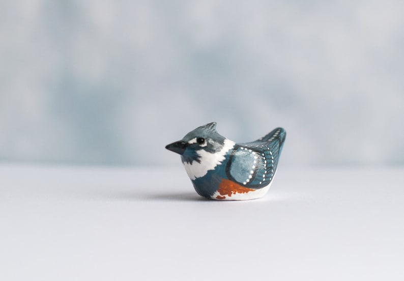 May include: Hand-painted ceramic bird figurine with blue, white, and orange details. The bird is perched on a white surface.