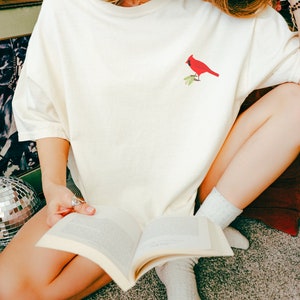 May include: A woman wearing a white t-shirt with a red cardinal embroidered on the front. She is sitting on a carpet and reading a book.
