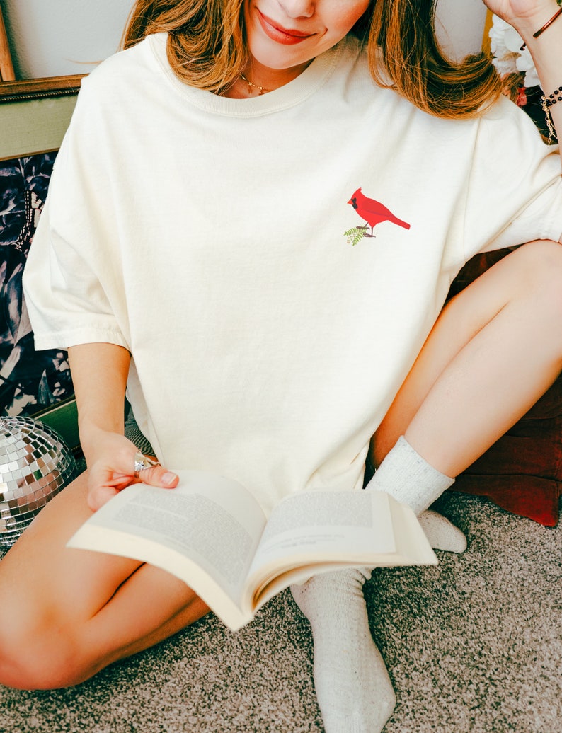 May include: A woman wearing a white t-shirt with a red cardinal embroidered on the front. She is sitting on a carpet and reading a book.