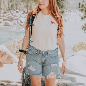 May include: A woman wearing a pale yellow t-shirt with a red cardinal embroidered on the front. She is also wearing blue denim shorts with distressed details and a blue and black rucksack.