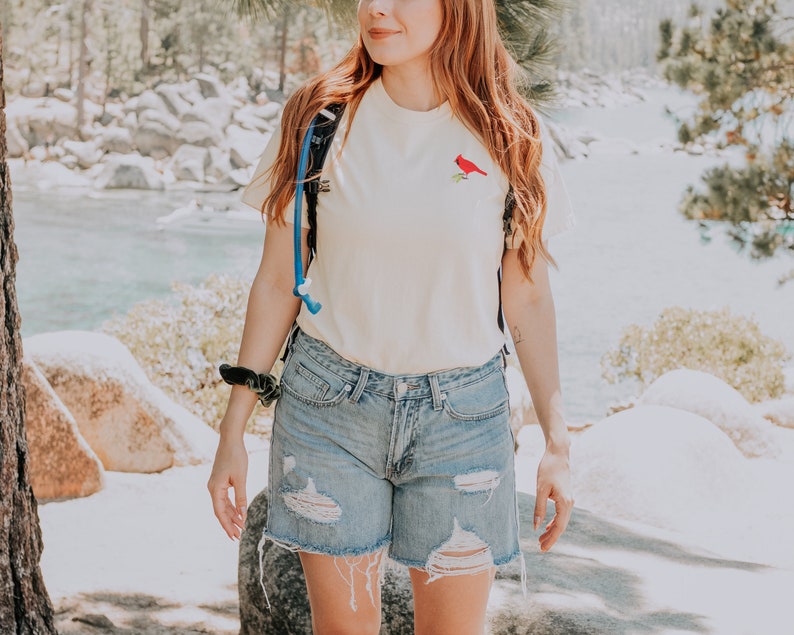 May include: A woman wearing a pale yellow t-shirt with a red cardinal embroidered on the front. She is also wearing blue denim shorts with distressed details and a blue and black rucksack.