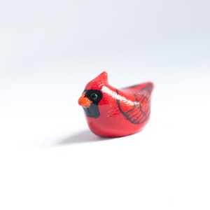 Peut inclure: Figurine de petit cardinal rouge avec des d&eacute;tails noirs et orange. L'oiseau est repr&eacute;sent&eacute; perch&eacute; et pr&eacute;sente des marques d'ailes d&eacute;taill&eacute;es. La figurine est pos&eacute;e sur un fond blanc, mettant en valeur ses couleurs vives et son design.
