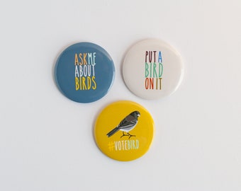 Pode incluir: Três botões redondos com designs diferentes. O primeiro botão é azul com texto branco que diz "Ask me about birds". O segundo botão é branco com texto colorido que diz "Put a bird on it". O terceiro botão é amarelo com uma ilustração de pássaro preto e branco e o texto "#votebird".