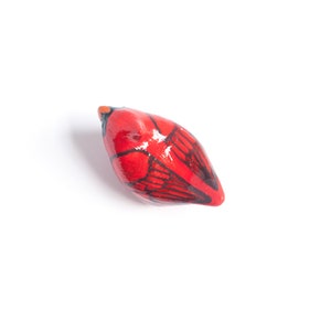 Peut inclure: Petite figurine d'oiseau en c&eacute;ramique rouge, avec des d&eacute;tails d'ailes noires et un bec orange. L'oiseau est peint &agrave; la main et a une finition brillante. L'objet est d&eacute;coratif, probablement pour l'exposition.
