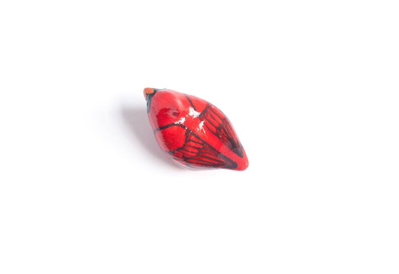 Peut inclure: Petite figurine d'oiseau en c&eacute;ramique rouge, avec des d&eacute;tails d'ailes noires et un bec orange. L'oiseau est peint &agrave; la main et a une finition brillante. L'objet est d&eacute;coratif, probablement pour l'exposition.