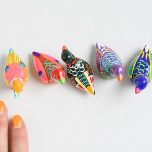 May include: Five colorful, hand-painted bird figurines in various colors, including yellow, orange, red, green, and purple. The birds are arranged on a white surface. The image also includes a hand with orange nail polish.