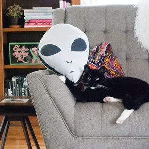 May include: A gray couch with a black cat and a white alien-shaped pillow. The couch has a colorful patterned throw pillow and a white fluffy pillow. The cat has white paws and a white patch on its chest.
